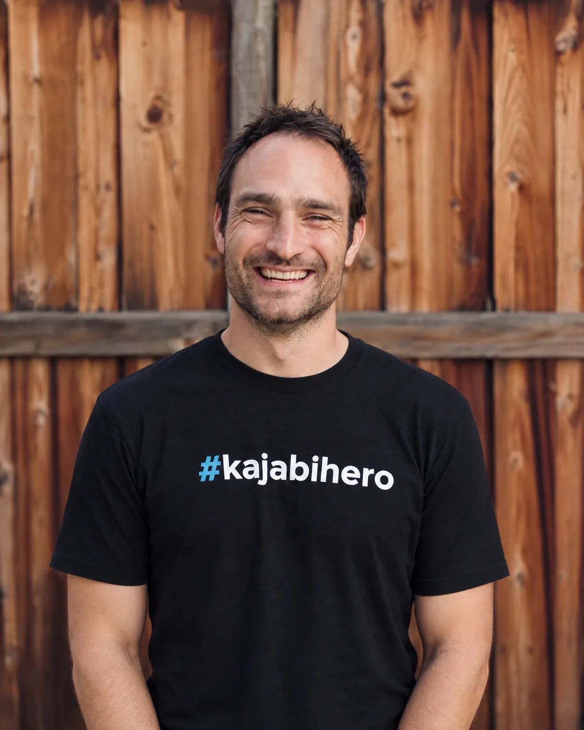 A smiling man in a black #kajabihero tee against a wooden fence