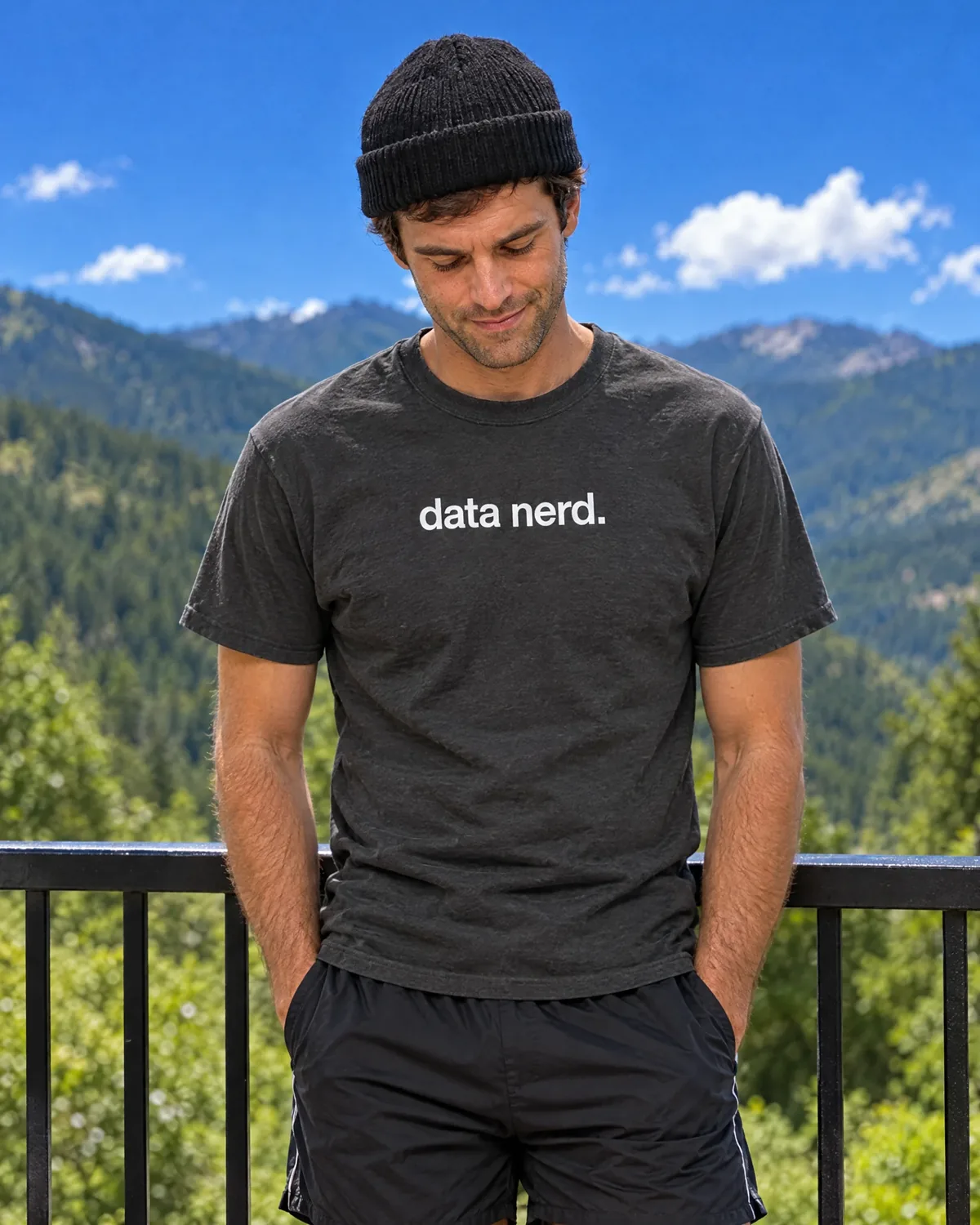 A man in a charcoal 'data nerd.' tee and black beanie on a balcony with mountains behind